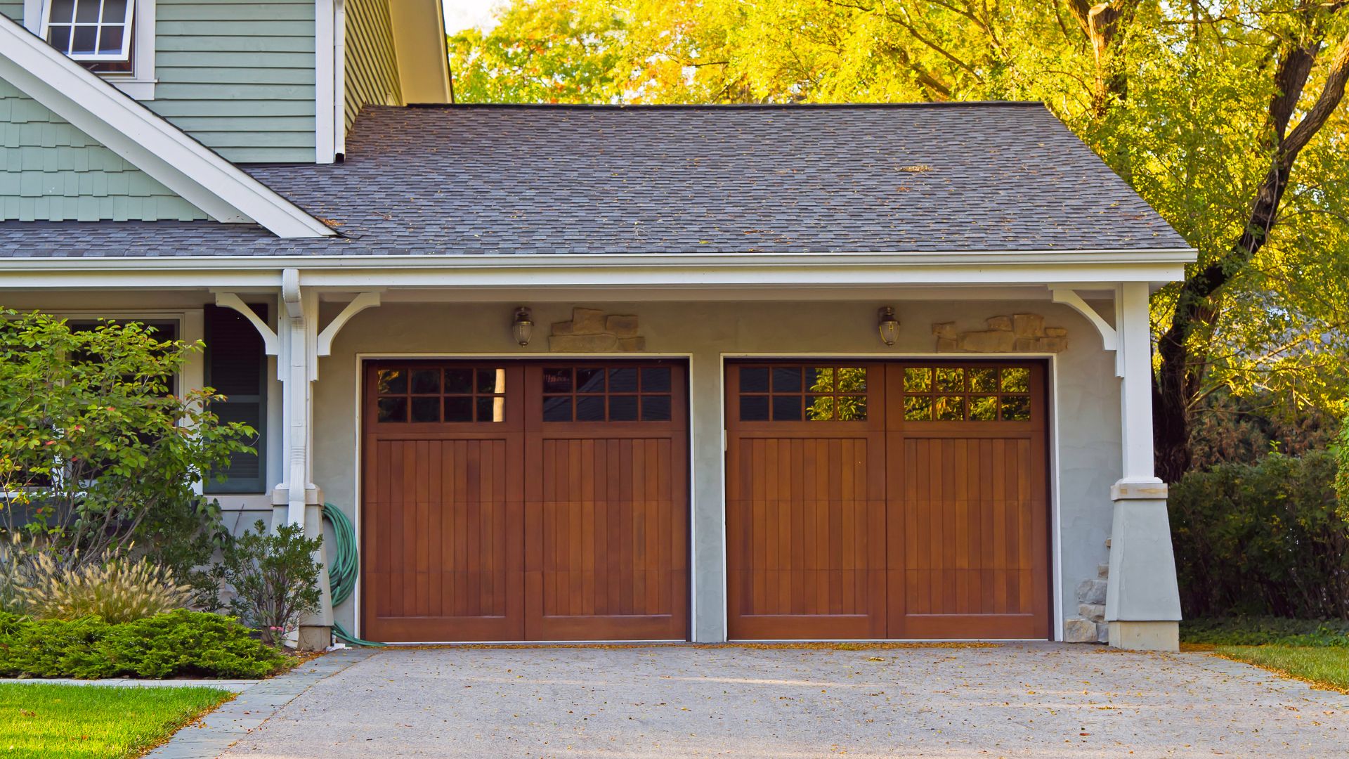 Two wooden garage doors on a green house with autumn trees in background